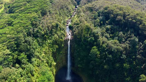 a drone lingers over a slender rainforest waterfall that threads through green cliffs into a circular, shadowed pool, mist drifting across dense canopy and tiered stream, calm yet forceful