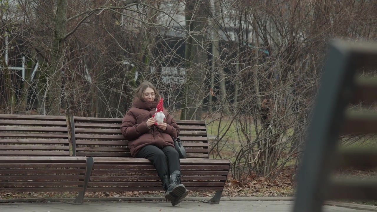White woman clutching red toy on park bench, hooded puffer coat, cold gray day, leafless trees and muted city backdrop, introspective posture, clutching memento, still, contemplative mood