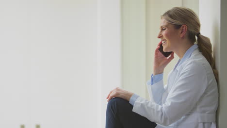 Female-Doctor-Wearing-White-Coat-Sitting-On-Floor-In-Hospital-Corridor-Using-Mobile-Phone