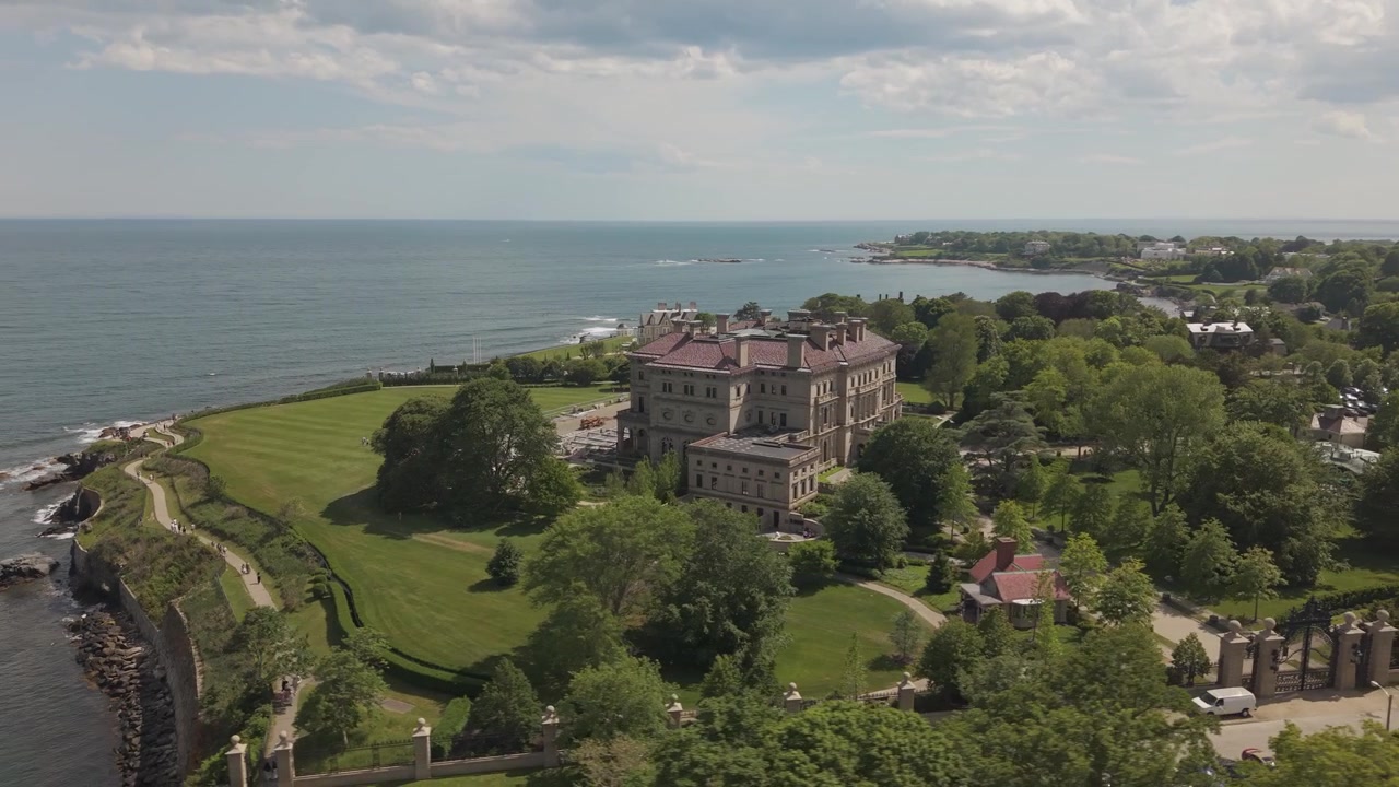 Herskapshuset The Breakers' storslåtte steinfasade reiser seg over en velstelt anlagt plen med utsikt over den steinete atlanterhavskysten i Newport, Rhode Island, under en delvis skyet himmel. Bredt dronebilde fra luften