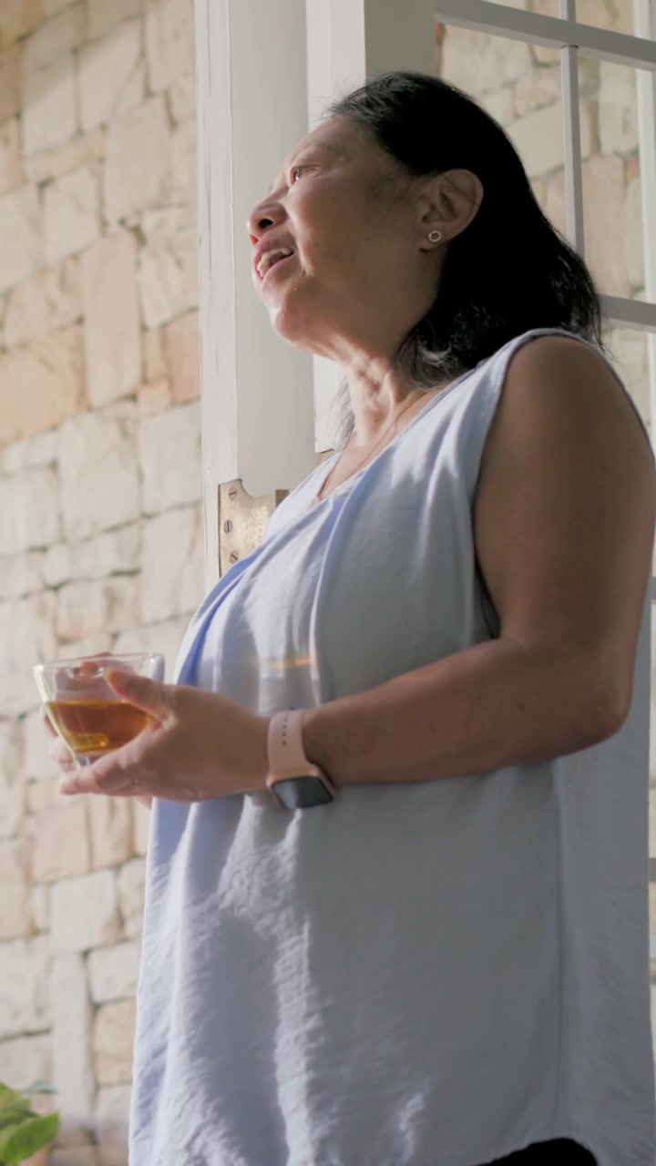 Video verticale: Donna anziana con top blu che entra e sorseggia pensierosa da una tazza di vetro alla finestra di casa.
