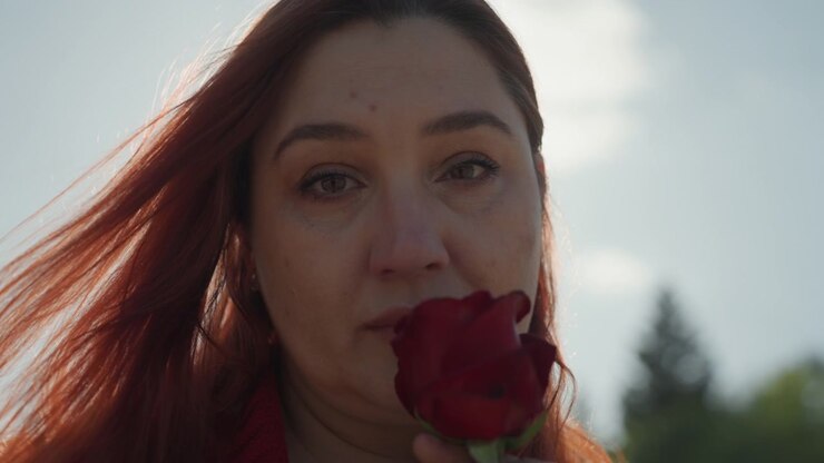 Close up of woman with long red hair holding and smelling single deep red rose outdoors under bright sunlight. Her expression is calm as woman gently brings flower to her face capturing soft