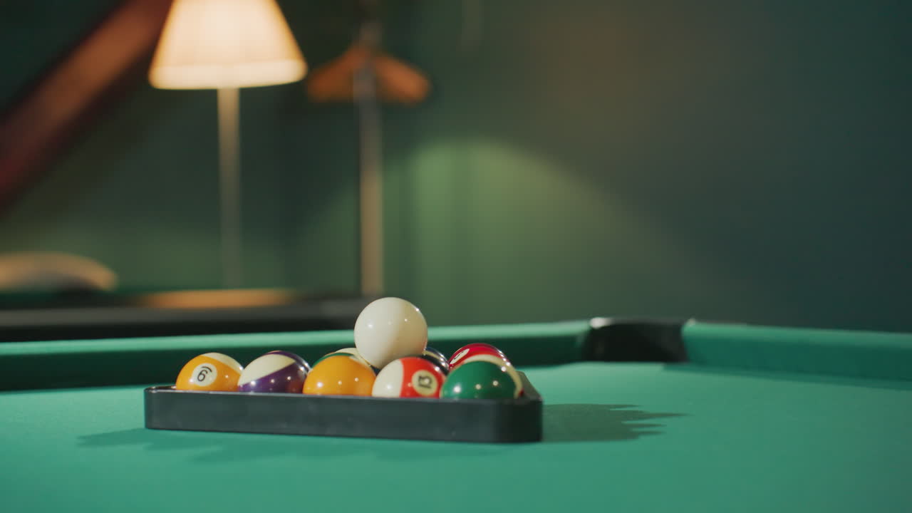 Close-up shot of colorful billiard balls arranged in triangle rack on green pool table as camera zooms in, capturing vibrant colors and smooth texture under soft warm lighting in cozy game room