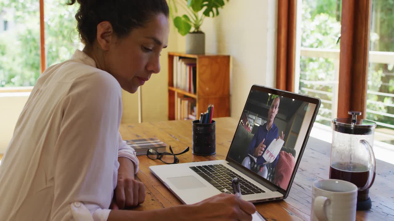 African american woman taking notes while having video call with male colleague on laptop at home