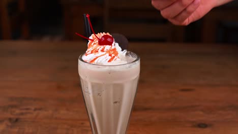 Detailed-close-up-of-chocolate-milkshake,-with-caramel,-espresso-coffee-and-whipped-cream-with-cherry,-hand-taking-whipped-cream-with-a-chocolate-cookie-and-ice-cream-with-caramel