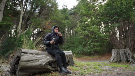 woman seated on log sipping yerba mate by snowy lakeshore in patagonia, argentina