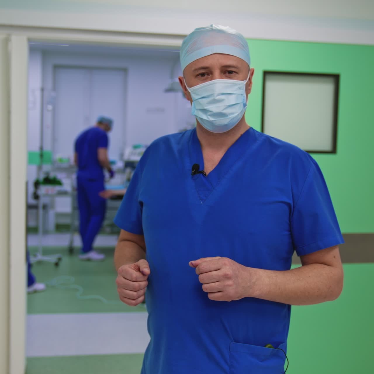 Portrait of a doctor in medical uniform and mask. Plastic surgeon speaking on camera in the hospital hall. Professional surgeon in operating room background