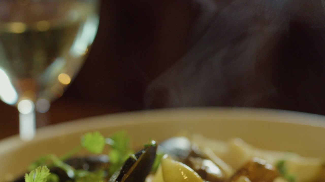 A close-up view of a hand scooping hot seafood chowder with visible steam, garnished with herbs, accompanied by a glass of white wine in warm indoor lighting