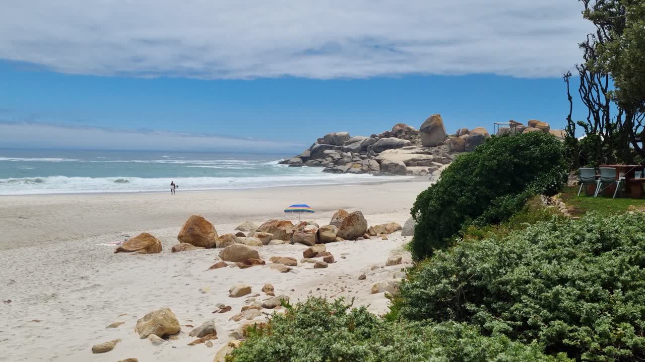 un surfista caminando en una playa de arena blanca de aspecto perfecto con agua azul y algunas formaciones rocosas en el fondo cerca de ciudad del cabo