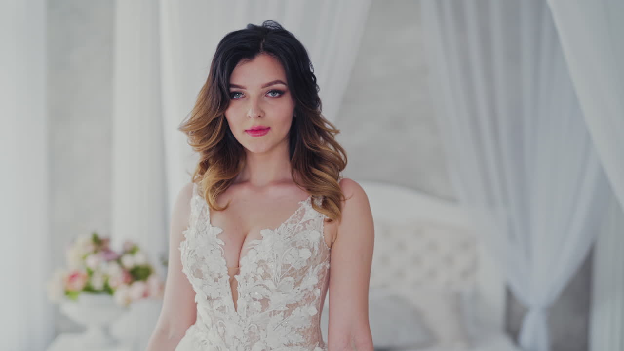 Sexy wedding lace dress on a beautiful model in the light studio. Attractive bride in luxurious dress with bouquet of flowers is coming to the window in bedroom.