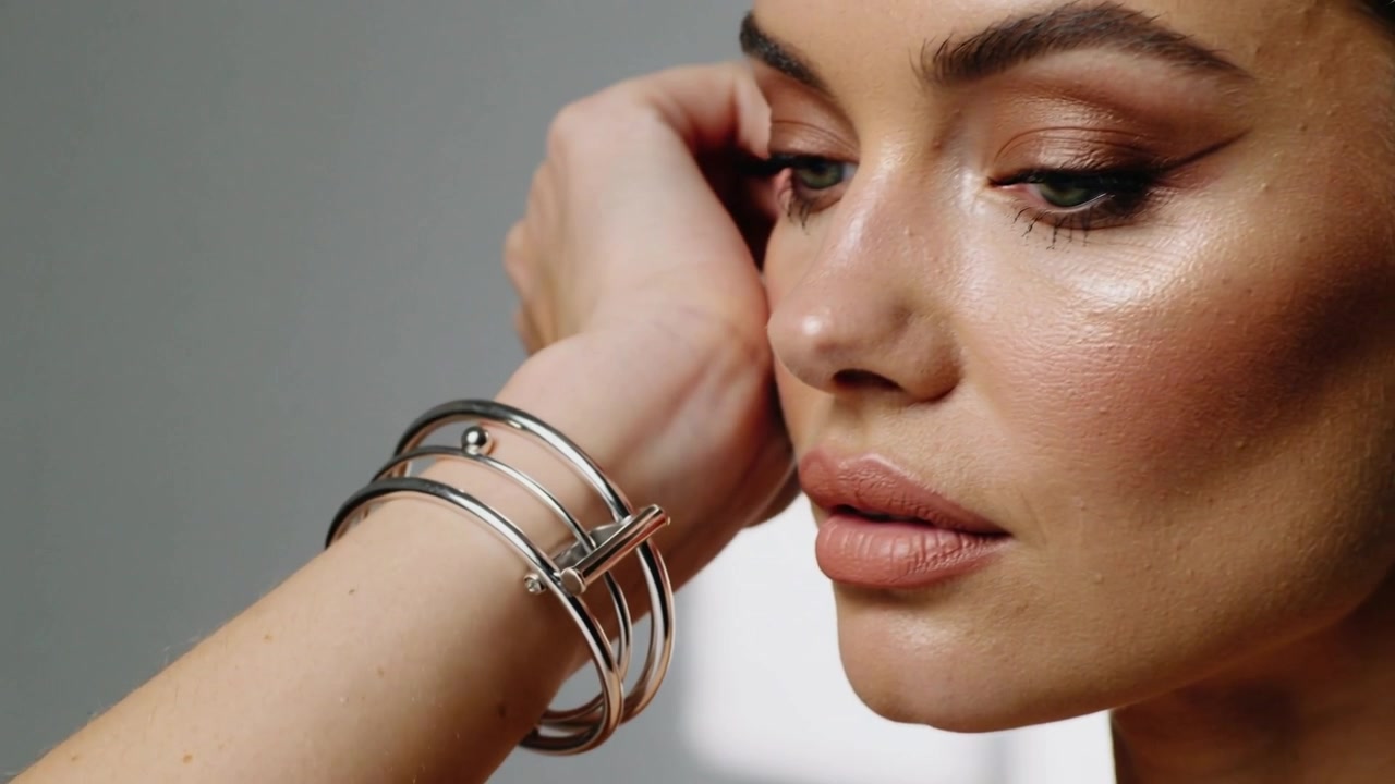 Lifting model tucking hair at temple in studio showing three linked silver bracelets, makeup for ad