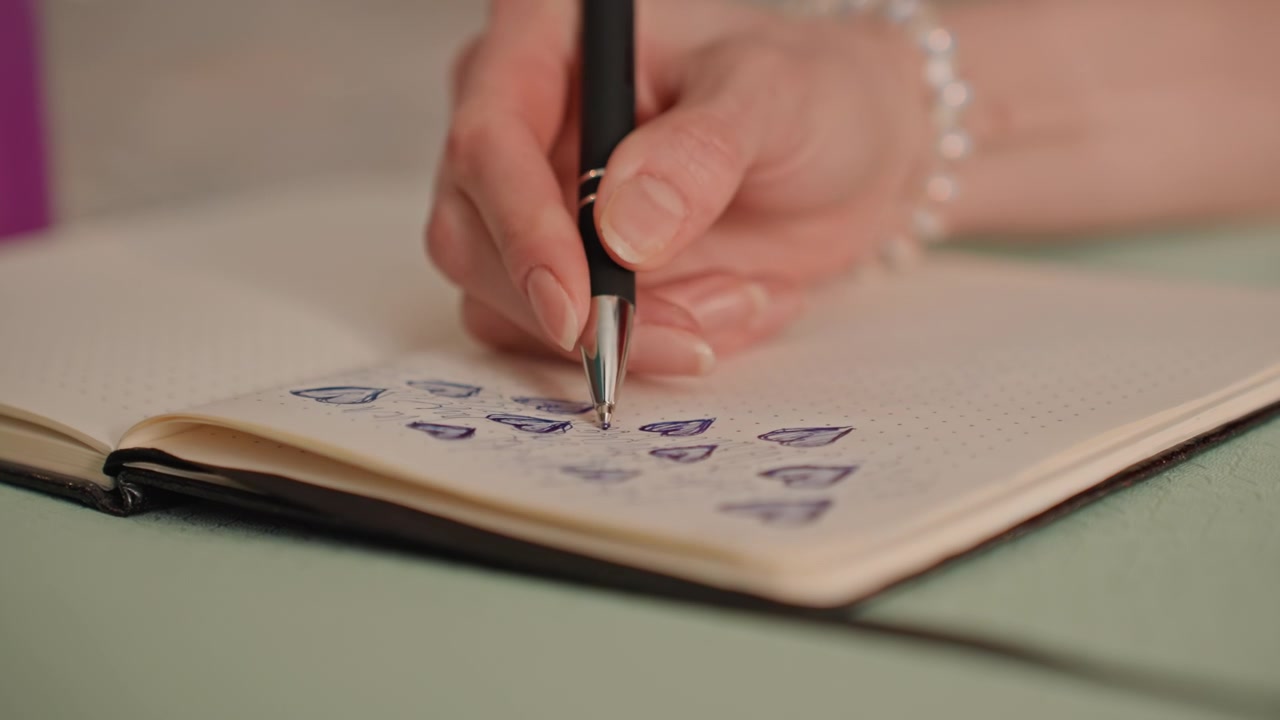 Weiße Frau zeichnet zu Hause Fitness-Tracker-Symbole in ein gepunktetes Notizbuch mit Stift und Perlenarmband, Nahaufnahme von Fingern und Tuschestrichen, sorgfältige Gewohnheitsplanung, ordentliches, organisiertes Layout und sanfter Nachmittag.