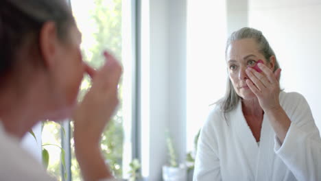 applying skincare, mature woman in bathrobe looking in mirror at home, focusing on face