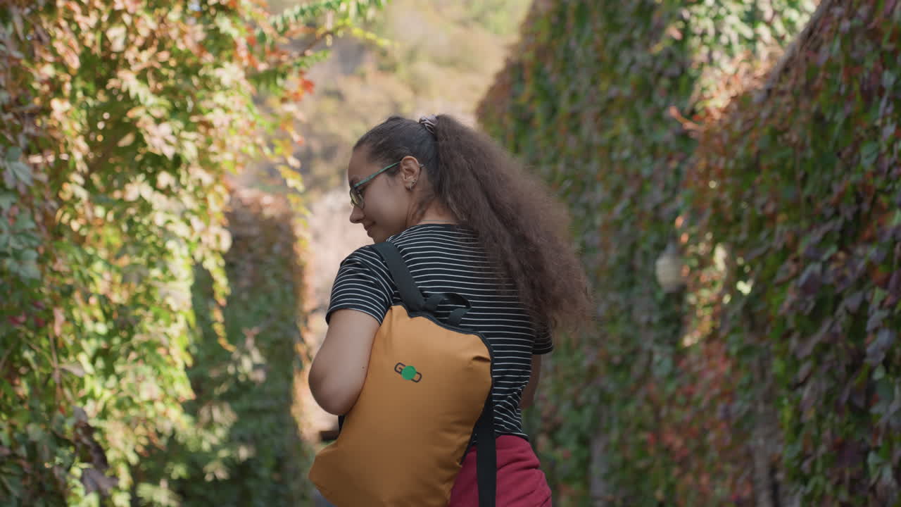 Woman leisurely explores vinecovered corridor. Young woman with backpack wanders along shaded ivy pathway. Young girl strolling casually through alleyway covered with lush vines and greenery