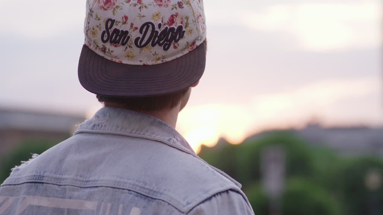 A slow motion shot of a young man with a cap looking into the camera and then turning around to watch the sunset