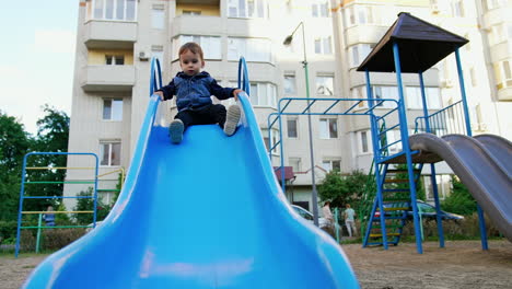 adorable toddler boy wearing jeans jacket sits on the top of a slide. kid waves his feet and then goes down. low angle view.