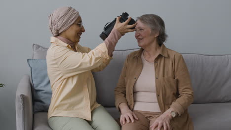 middle aged arabic woman helping a senior lady to use virtual reality headset glasses at home