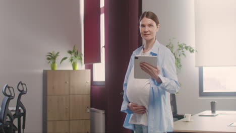 Mujer-Embarazada-Posando-En-La-Cámara-Mientras-Toca-Su-Vientre-Y-Sostiene-Una-Tableta-En-La-Oficina