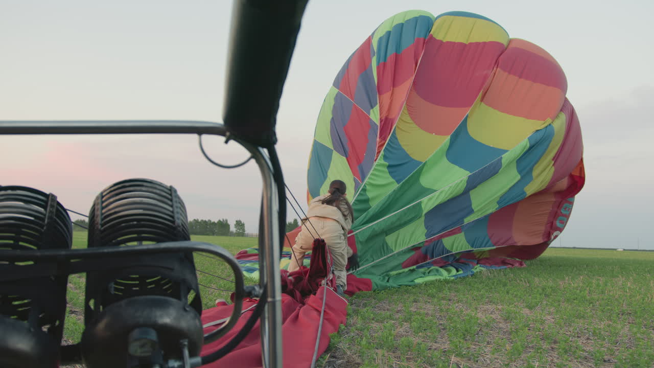 back view of woman in beige uniform folding colorful hot air balloon on grassy field beside metal burner frame under soft evening light