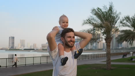 Loving-Father-in-a-white-t-shirt-in-the-summer-walks-with-a-child-sitting-on-the-neck-against-the-city-and-palm-trees