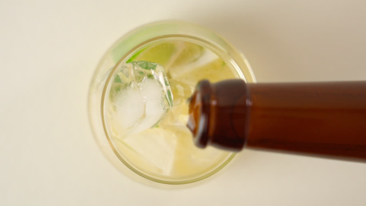 Ripresa dall'alto di una bottiglia di birra che versa una bevanda in un bicchiere pieno di ghiaccio e fette di lime a 100 FPS su sfondo bianco