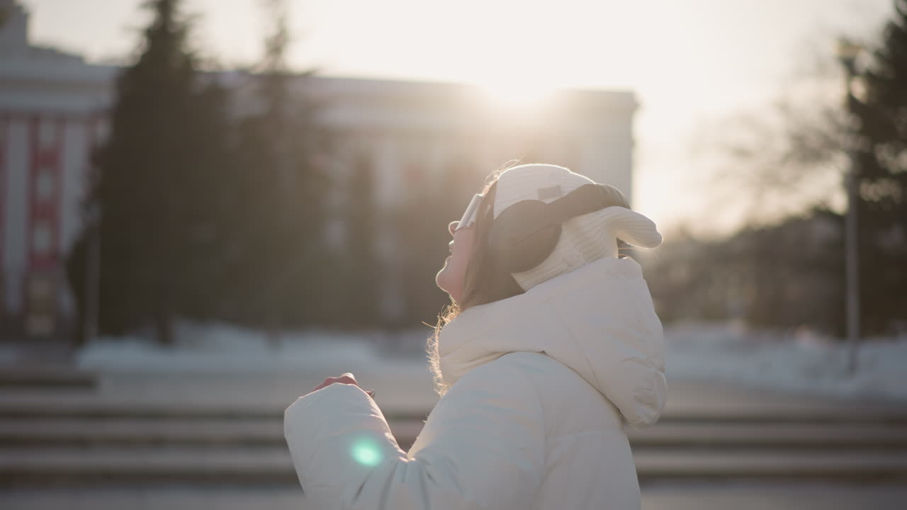 Young lady dances with rhythm in winter outfit, surrounded by tall buildings, as warm sunset light highlights her joyful expression, headphones on and full of energy in urban atmosphere