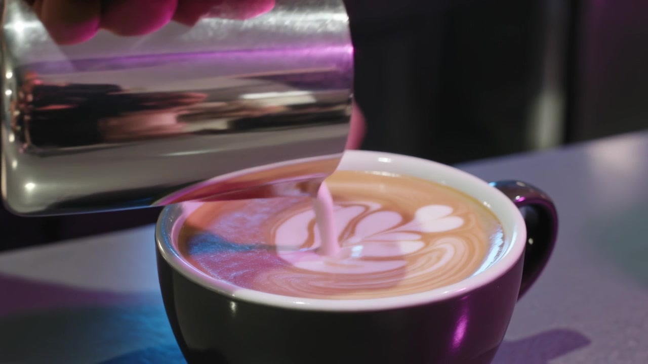 Inclinando una jarra metálica, una mano con manga vertiendo leche en una taza negra en una cafetería, formando una roseta, sirviendo.