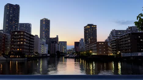 Skyscrapers-and-cityscape-at-dusk-reflect-on-a-calm-river-under-a-twilight-sky