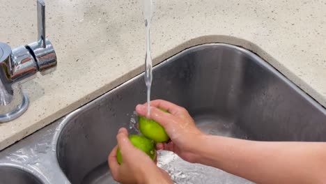 hands thoroughly rinse green limes under a steady stream of water in a modern kitchen sink, with bright, even lighting and close-up framing