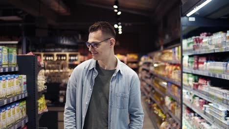 A-handsome-young-caucasian-guy-in-glasses-and-blue-shirt-doing-shopping-in-the-supermarket,-walking-by-aisle,-looks-to-the-sides-and-smiling-cheerfully.-Slow-motion