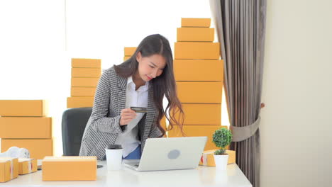 asian woman received big payment on card balance doing an online sales with her laptop computer, courier parcel boxes at the home office