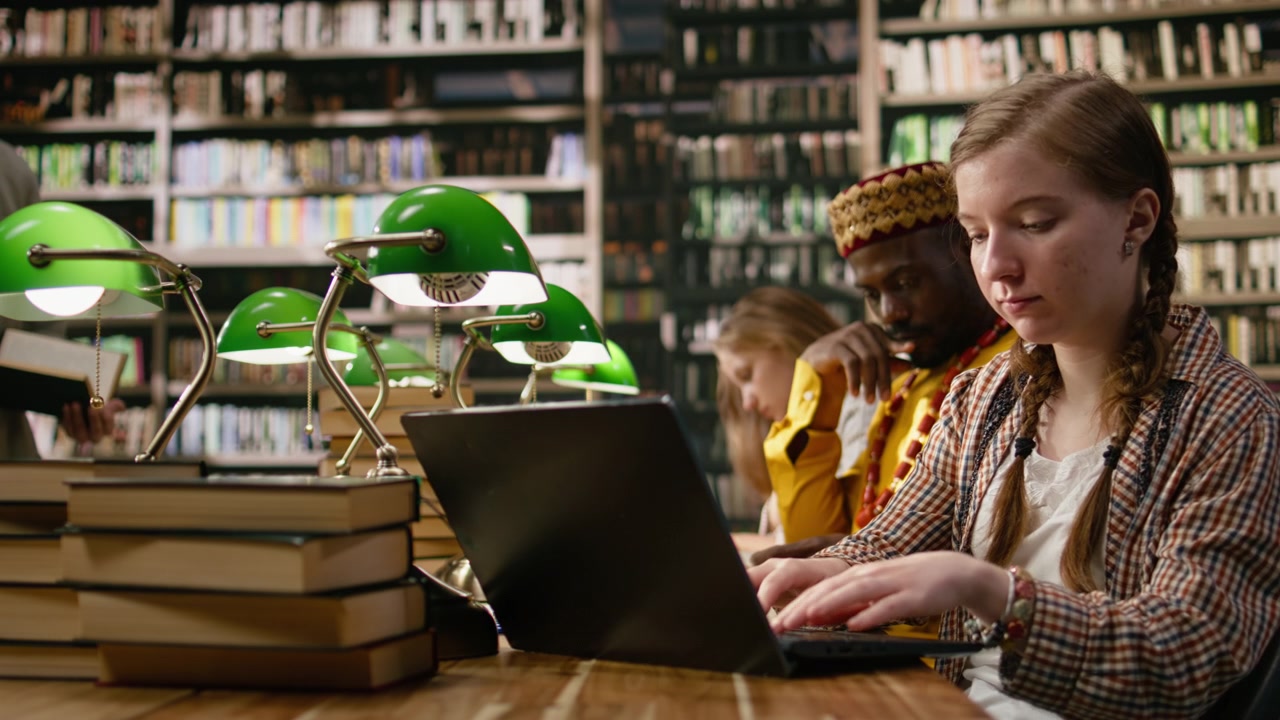 Student typing on laptop and taking notes beside open book at busy study table