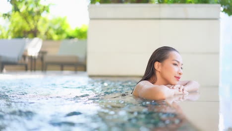 an attractive young woman moves through the pool water to the edge of the pool then rests her arms and head against the edge