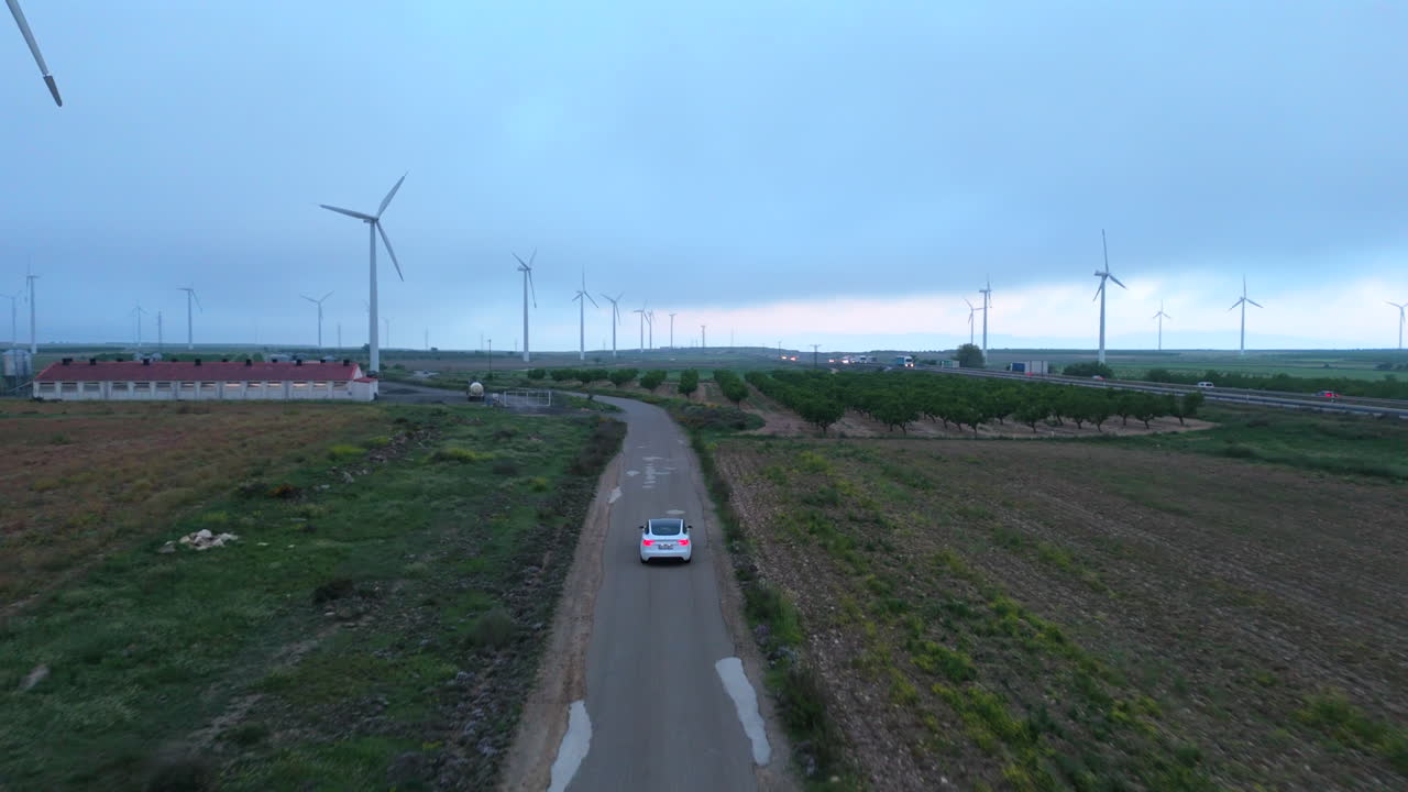 Electric car driving between wind turbines at sunrise, followed from behind by a drone flying low over rural roads and agricultural fields