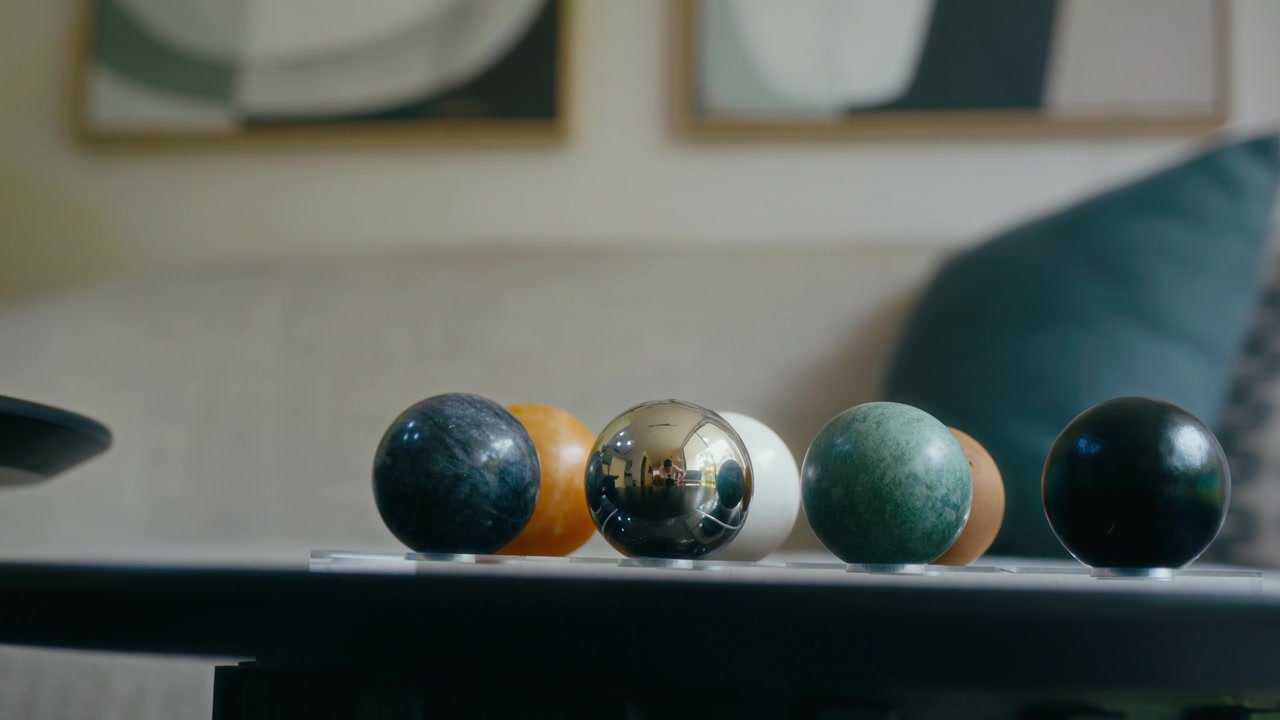 Close-up of five colorful spheres on black table. Smooth surfaces reflect light softly. Table sits near sofa with cushions. Wall art features abstract patterns. Modern living room scene