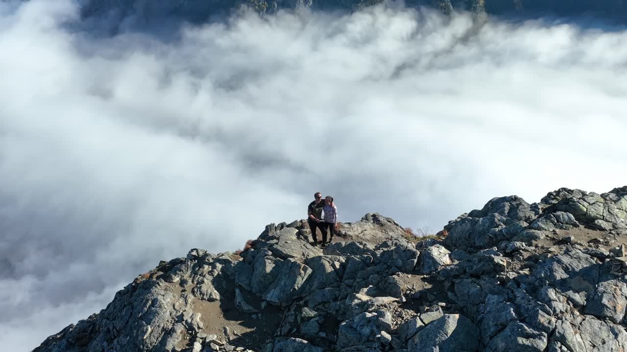 Drone orbits and zooms in on a couple at Bakkanosi cliffs, creating a parallax effect with the distant fjord below