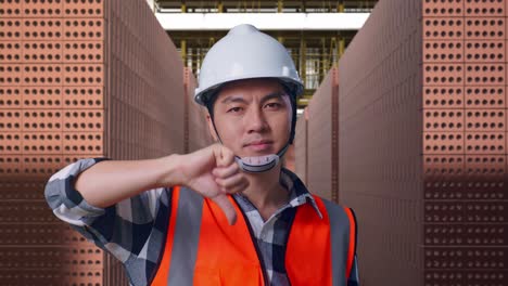 close up of asian male engineer with safety helmet showing thumbs down gesture and shaking his head while standing with red brick packed in stacks are stored