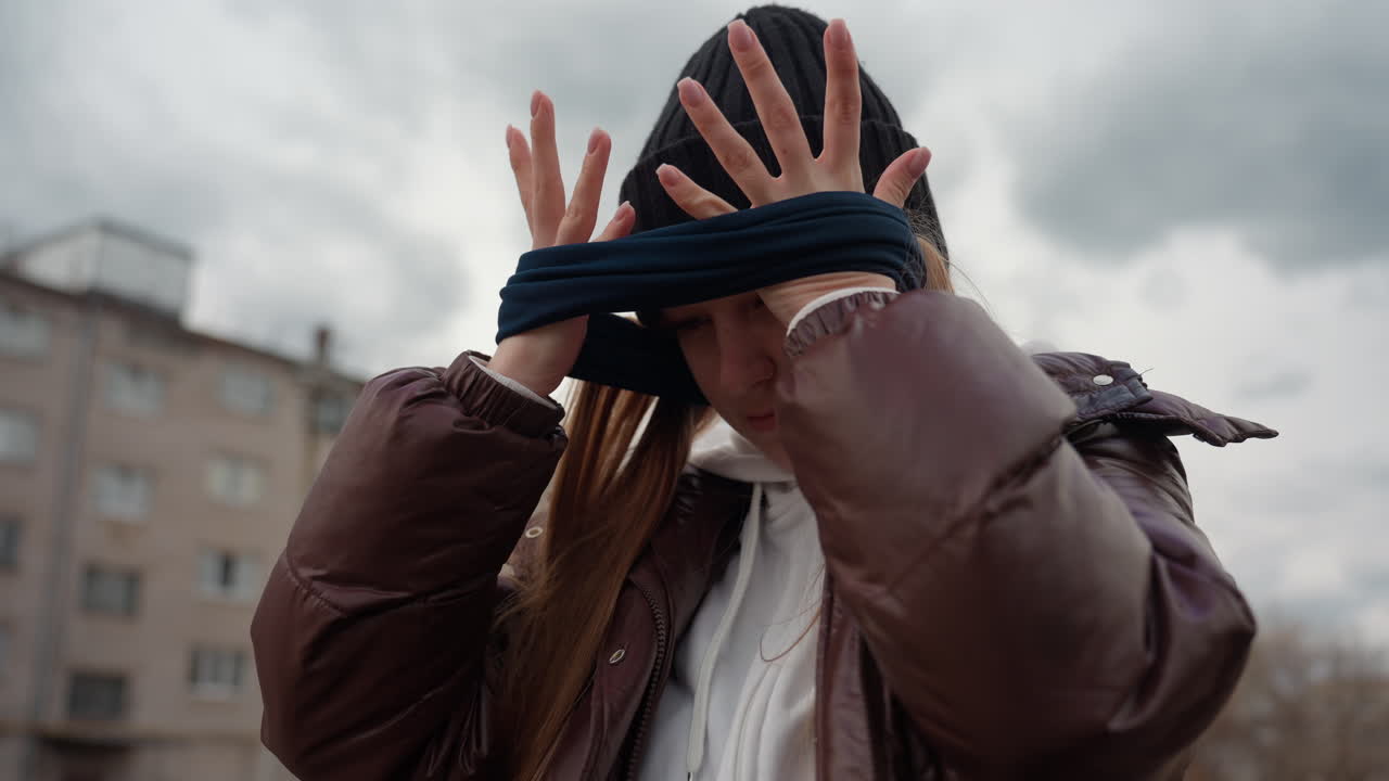 Mujer caucásica ajustándose la bufanda por el cuello en una calle nublada de la ciudad, gorro, chaqueta acolchada, pelo largo, manos alisando tejido de punto, expresión serena, edificios universitarios, cielo nublado, trayecto invernal