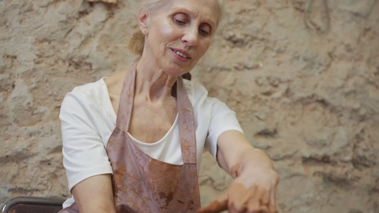 Old woman potter working on Potter wheel making a clay pot. Master forming the clay with her hands creating pot in a workshop