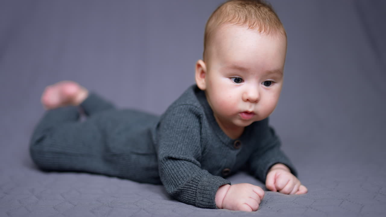 Sweet Caucasian child in grey outfit lies on belly on the bed. Beautiful child looking around with surprise. Grey backdrop.