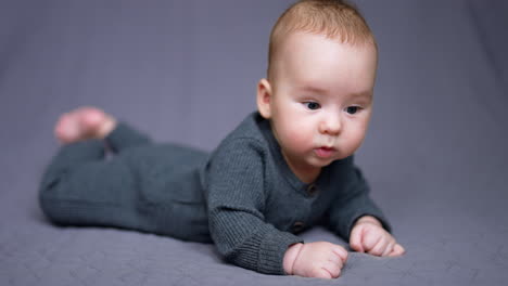 sweet caucasian child in grey outfit lies on belly on the bed. beautiful child looking around with surprise. grey backdrop.