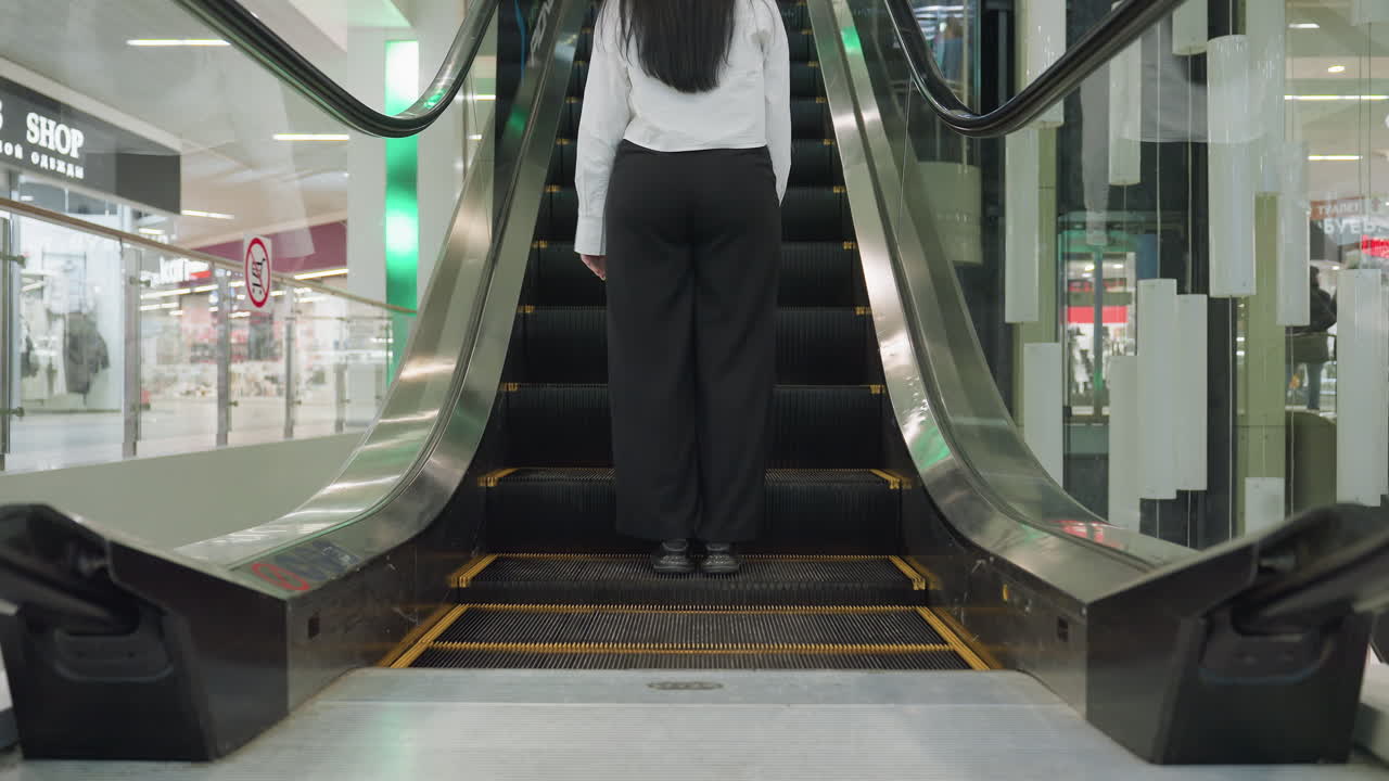 Back view of woman wearing black pants and white shirt stepping onto moving escalator inside brightly lit modern shopping mall with reflections, glass panels, and retail stores in background