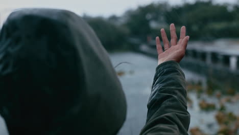 young woman with arms raised standing by lake on cloudy day catching rain in hands with wind blowing hair