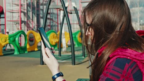 brunette woman using smartphone and watching children playing on playground