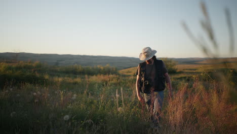 man with hat and camera walks through field of grass and wildflowers