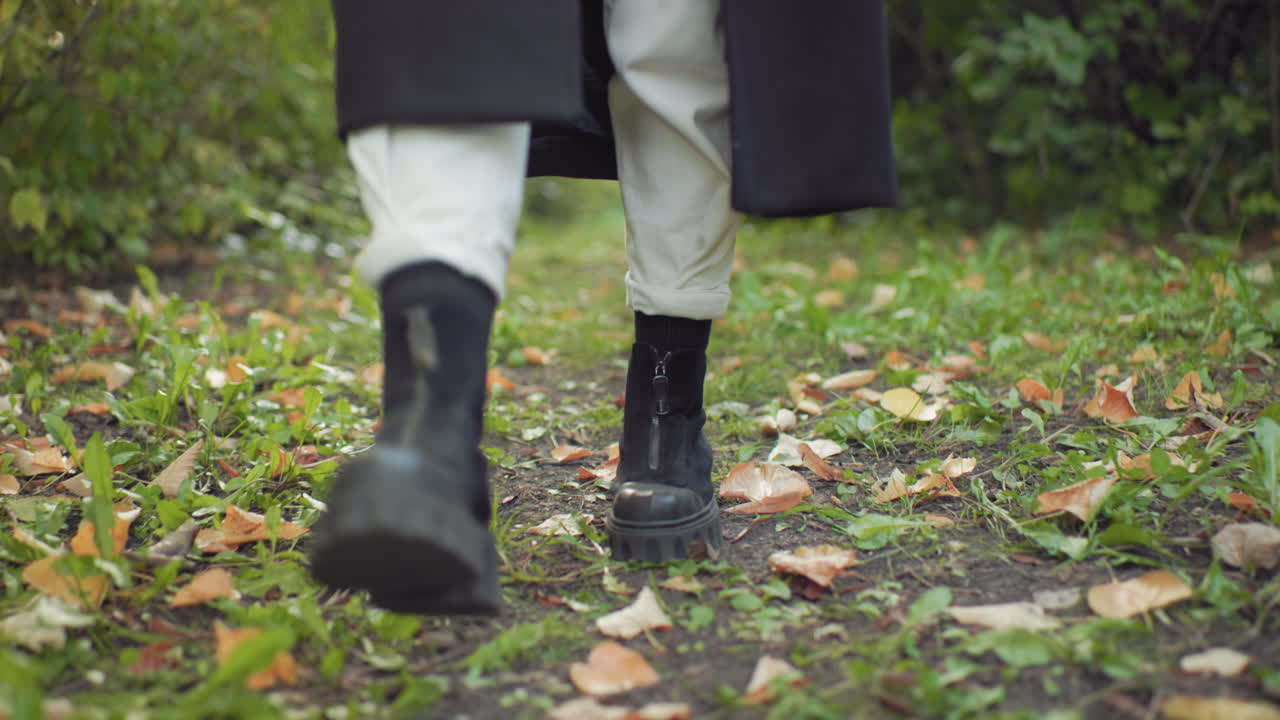 Leg view of autumn woman walking in black boots along green forest path, coat hem swaying, steps over scattered leaves, calm pace under soft light, fresh foliage around, seasonal mood of quiet travel
