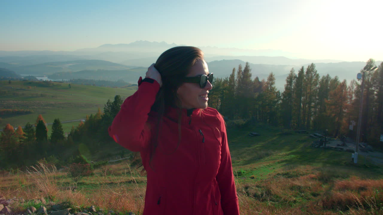 girl standing and smiling in a red jacket on a top a hill on a sunny day fixing her hair in slow motion