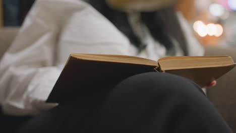 woman in white top seated with long black hair kneels in lounge flipping pages of book on her lap under soft evening light with bokeh background