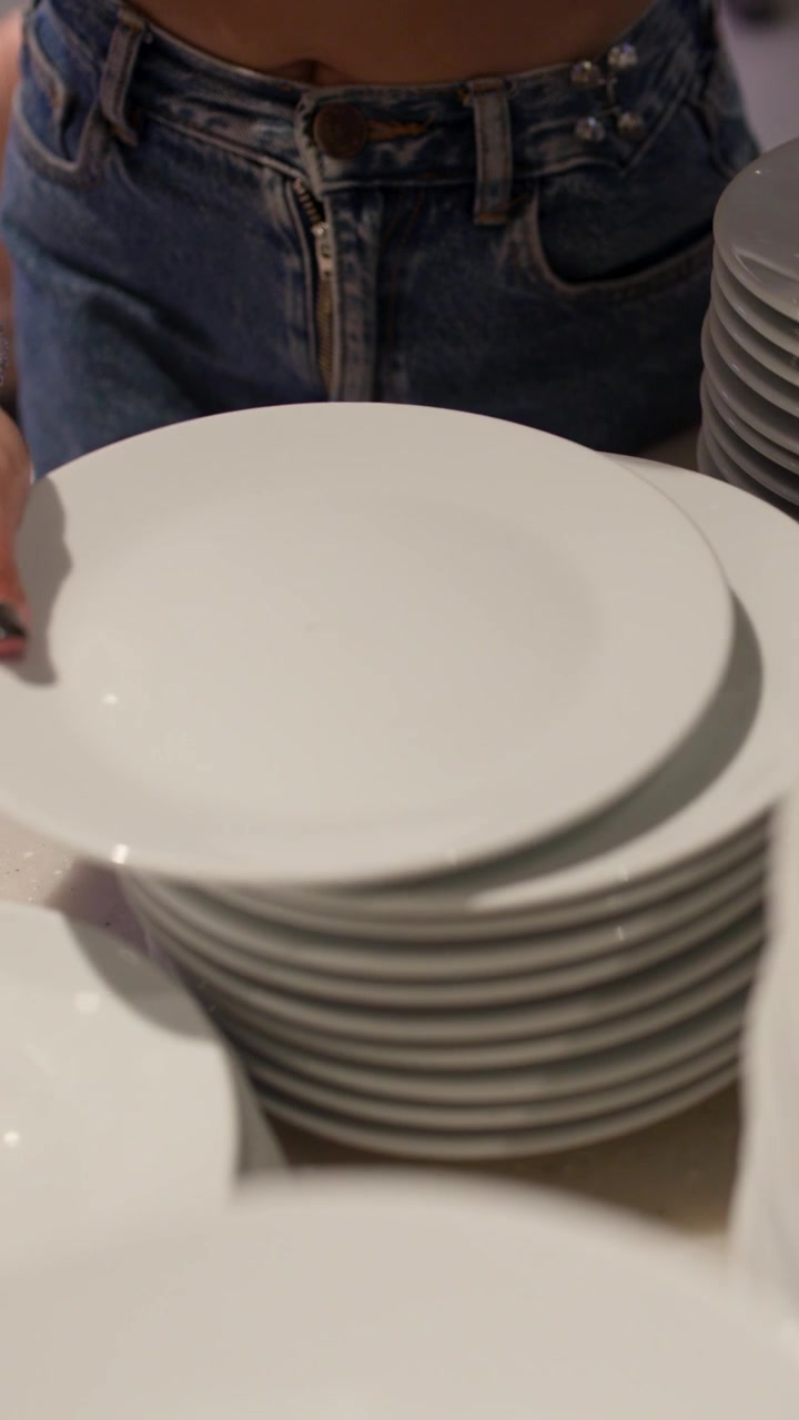 Close-up of woman arranging stacked white plates on buffet counter in softly lit restaurant