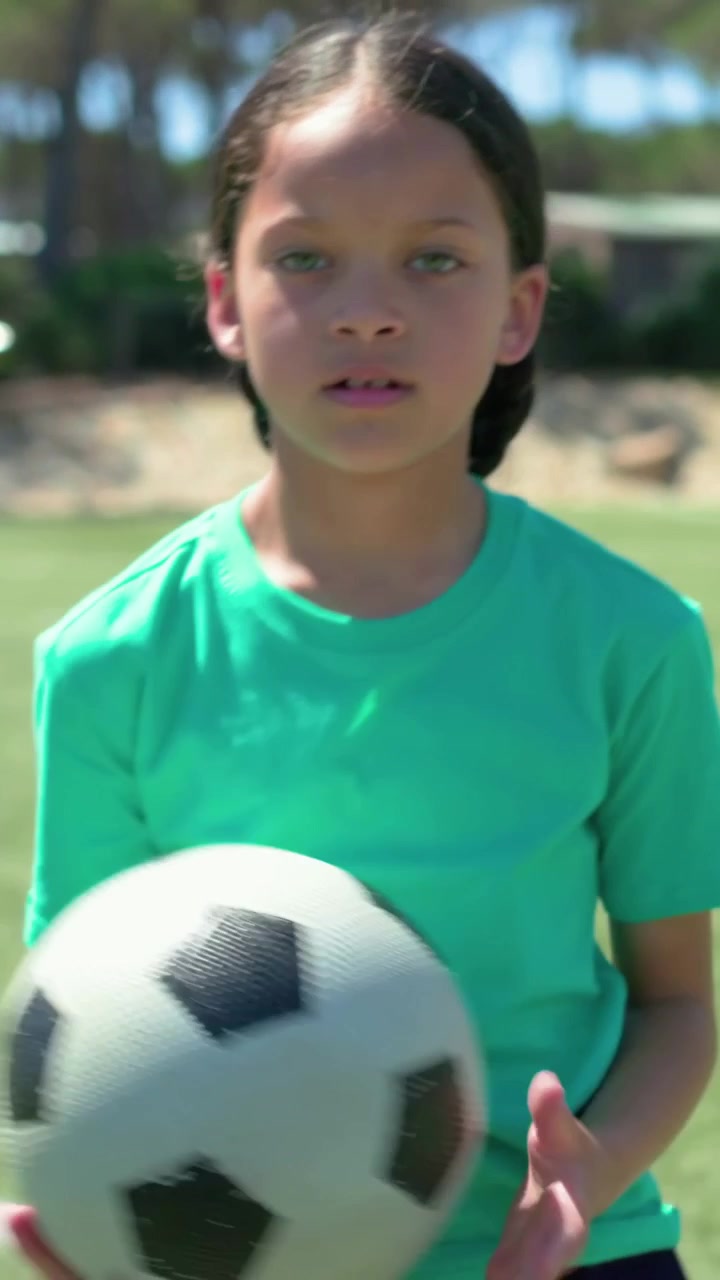 Vídeo vertical: Clipe focado na garota central de camisa verde fazendo embaixadinhas com uma bola de futebol em campo, praticando